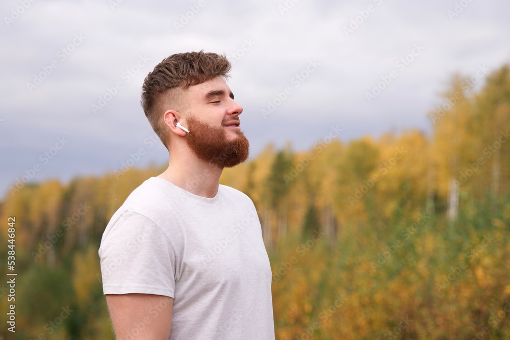 Young happy calm relaxed bearded man with beard enjoying listen to music in wireless earphones in the forest, park, breathing deep inhale fresh air and smiling with beard on natural background