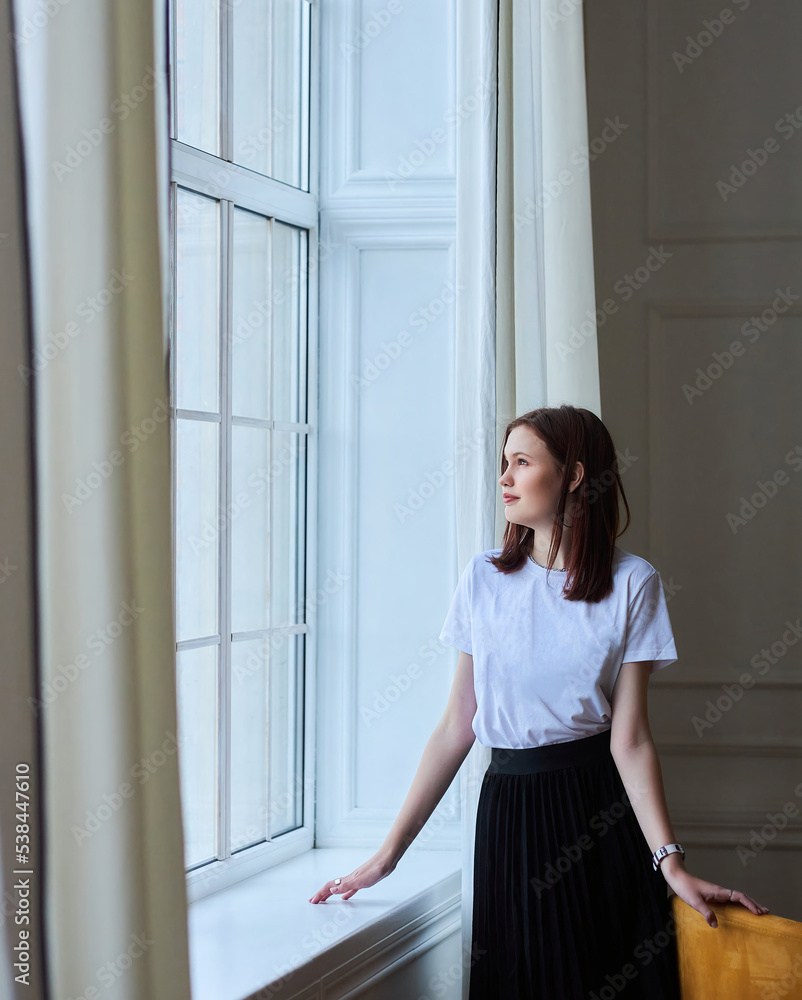 A young stylish girl is standing at the window in a bright room. Style and fashion. A young model. A young stylish girl is standing at the window in a bright room. Style and fashion. A young model.