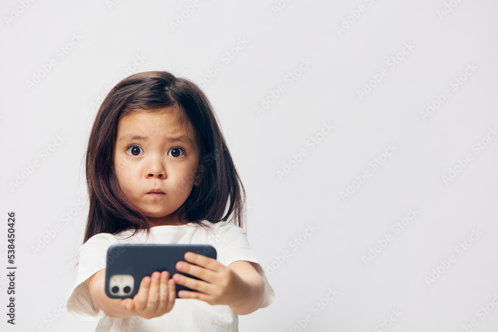 cute, funny, little girl in a white t-shirt stands on a light background with a smartphone in her hand and takes a selfie. Horizontal studio photography with blank space for advertising mockup insert