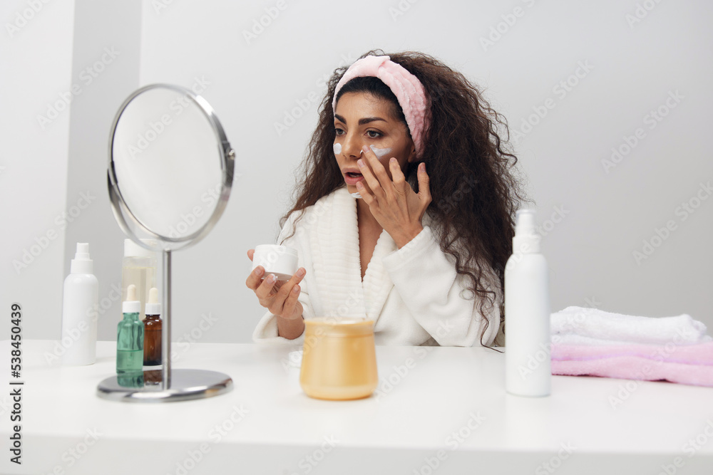 Flawless cute happy tanned curly Latin lady put facial cream on cheek area admiring herself smiling in mirror at home interior. Copy space. Aesthetic medicine skincare beauty products ad concept Flawless cute happy tanned curly Latin lady put facial cream on cheek area admiring herself smiling in mirror at home interior. Copy space. Aesthetic medicine skincare beauty products ad concept