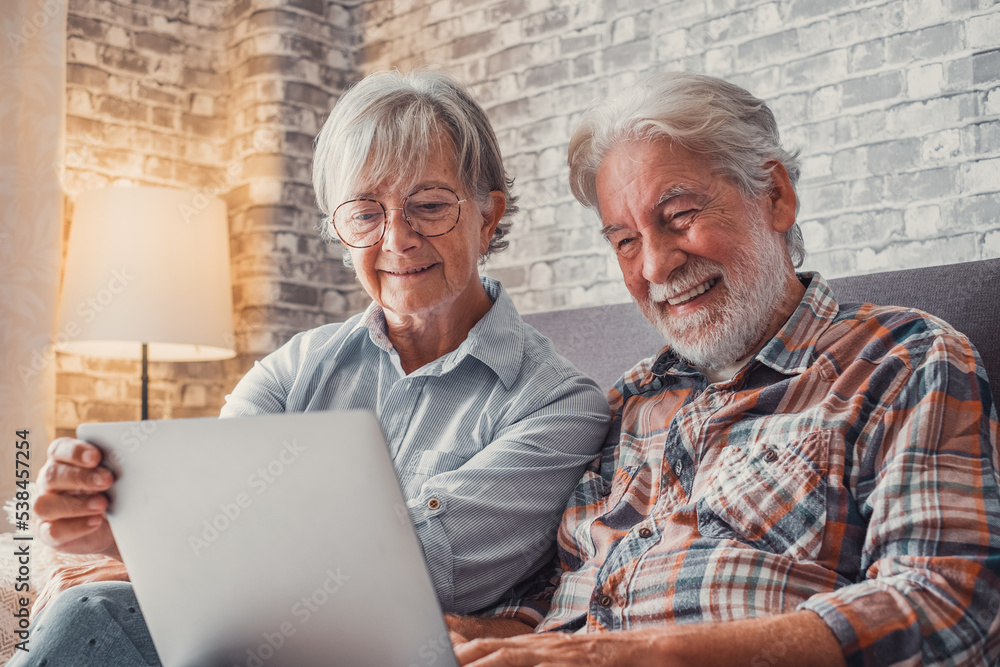 Cute couple of old people sitting on the sofa using laptop together shopping and surfing the net. Two mature people in the living room enjoying technology. Cute couple of old people sitting on the sofa using laptop together shopping and surfing the net. Two mature people in the living room enjoying technology.