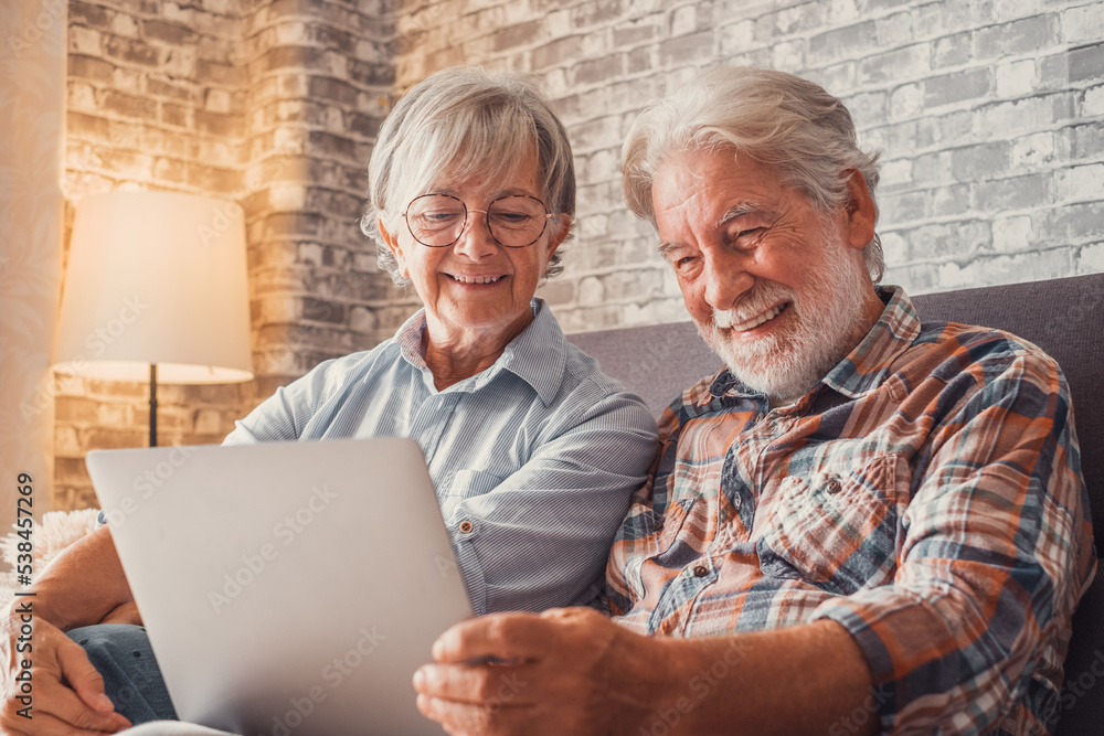 Cute couple of old people sitting on the sofa using laptop together shopping and surfing the net. Two mature people in the living room enjoying technology. Cute couple of old people sitting on the sofa using laptop together shopping and surfing the net. Two mature people in the living room enjoying technology.