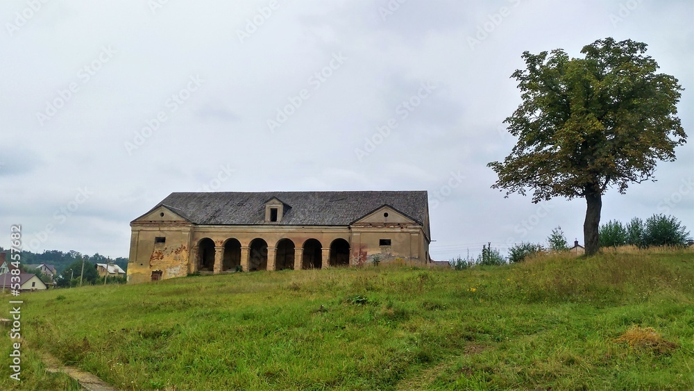 On the slope of a grassy hill there is a historical outbuilding with an arched colonnade and a wooden plank roof and a large tree grows. There is a cottage village behind the hill