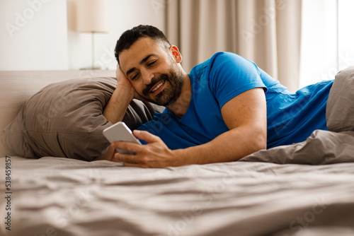 Communication and man concept - happy smiling man texting on smartphone in bed at home at night