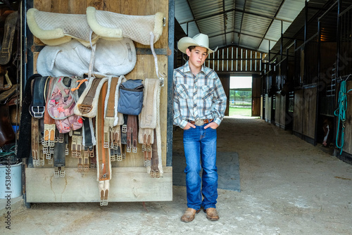 Young cowboy standing in a horse barn with saddles and other riding equipment stowed nearby