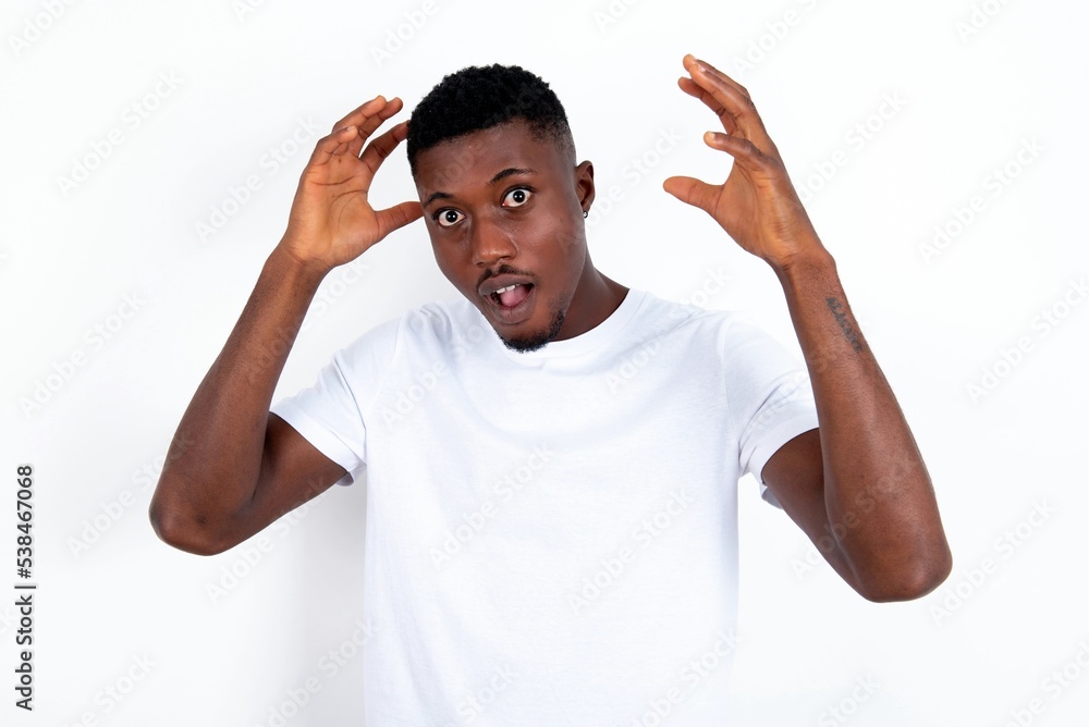 Fototapeta premium young handsome man wearing white T-shirt over white background looks with excitement at camera, keeps hands raised over head, notices something unexpected, reacts on sudden news.
