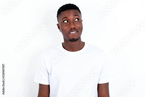 young handsome man wearing white T-shirt over white background with thoughtful expression, looks away keeps hands down bitting his lip thinks about something pleasant.