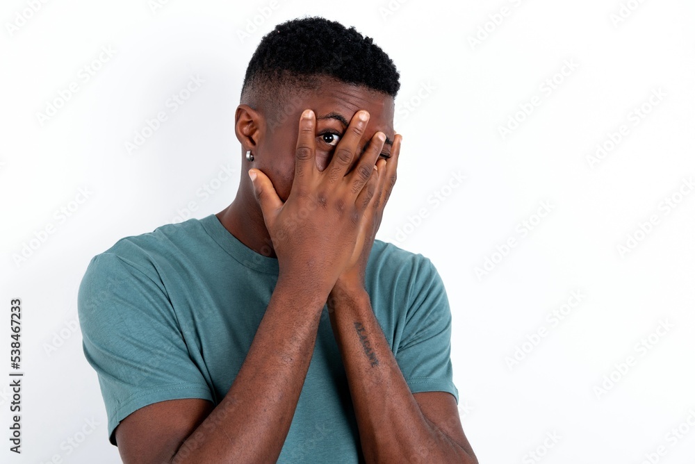 young handsome man wearing green T-shirt over white background covering face with hands and peering out with one eye between fingers. Scared from something or someone. young handsome man wearing green T-shirt over white background covering face with hands and peering out with one eye between fingers. Scared from something or someone.
