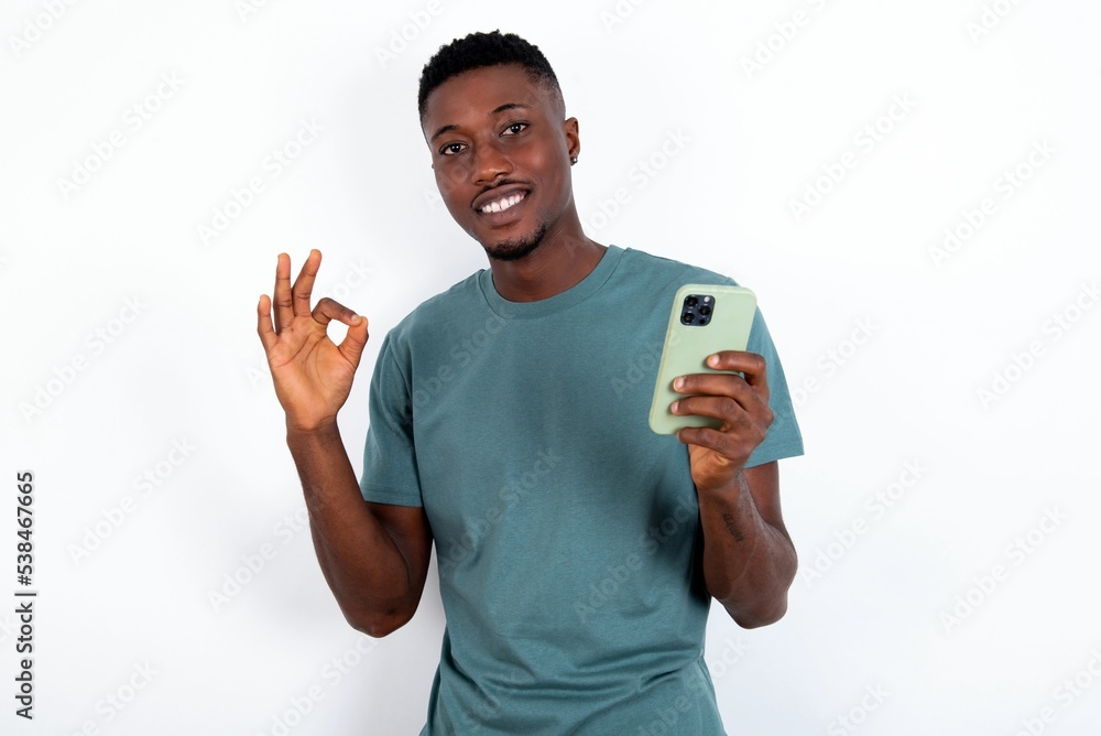 Happy young handsome man wearing green T-shirt over white background sending a message on his smartphone or taking a selfie and making ok sign with his hand. Happy young handsome man wearing green T-shirt over white background sending a message on his smartphone or taking a selfie and making ok sign with his hand.