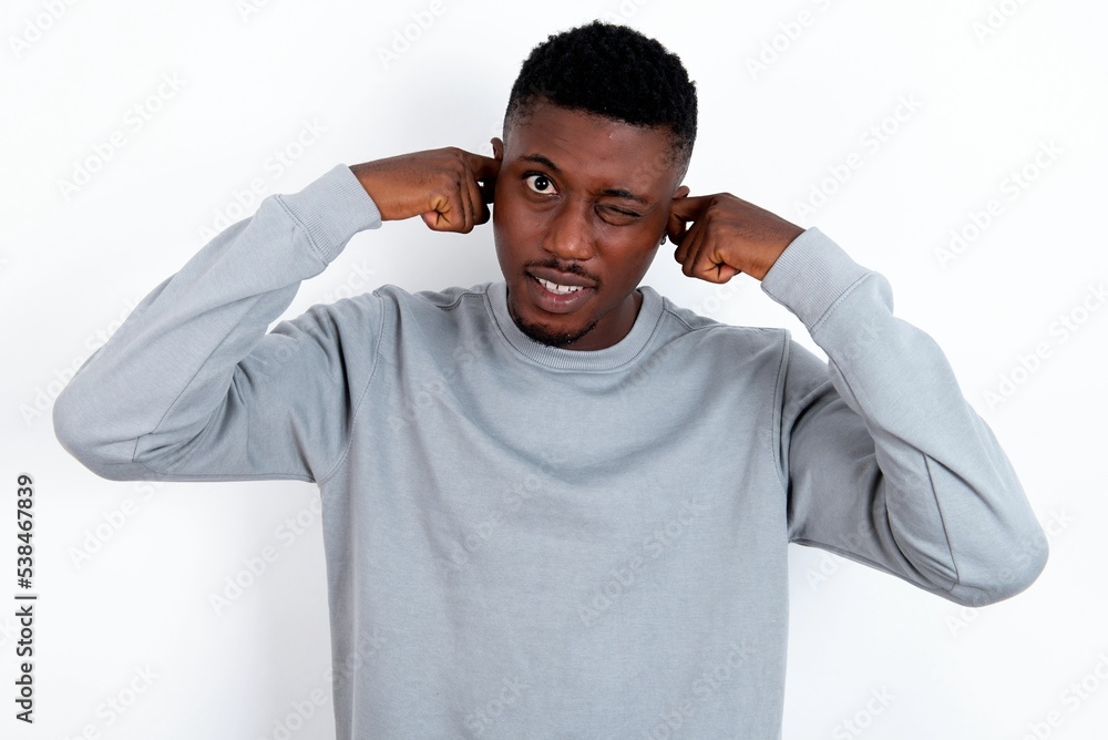 Happy young handsome man wearing grey sweater over white background ignores loud music and plugs ears with fingers asks to turn off sound Happy young handsome man wearing grey sweater over white background ignores loud music and plugs ears with fingers asks to turn off sound