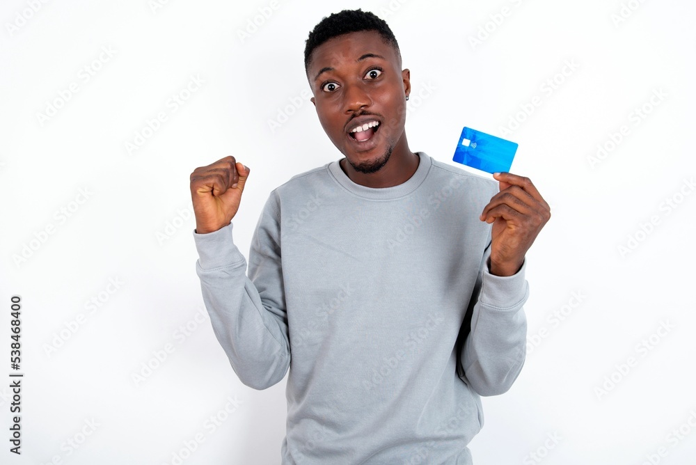 Photo of lucky impressed young handsome man wearing grey sweater over white background arm fist holding credit card. Celebrated Photo of lucky impressed young handsome man wearing grey sweater over white background arm fist holding credit card. Celebrated