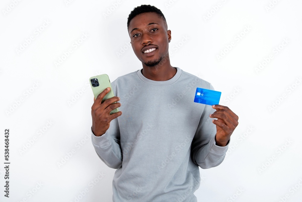 young handsome man wearing grey sweater over white background wears stereo headphones on ears holds modern mobile phone and credit card young handsome man wearing grey sweater over white background wears stereo headphones on ears holds modern mobile phone and credit card