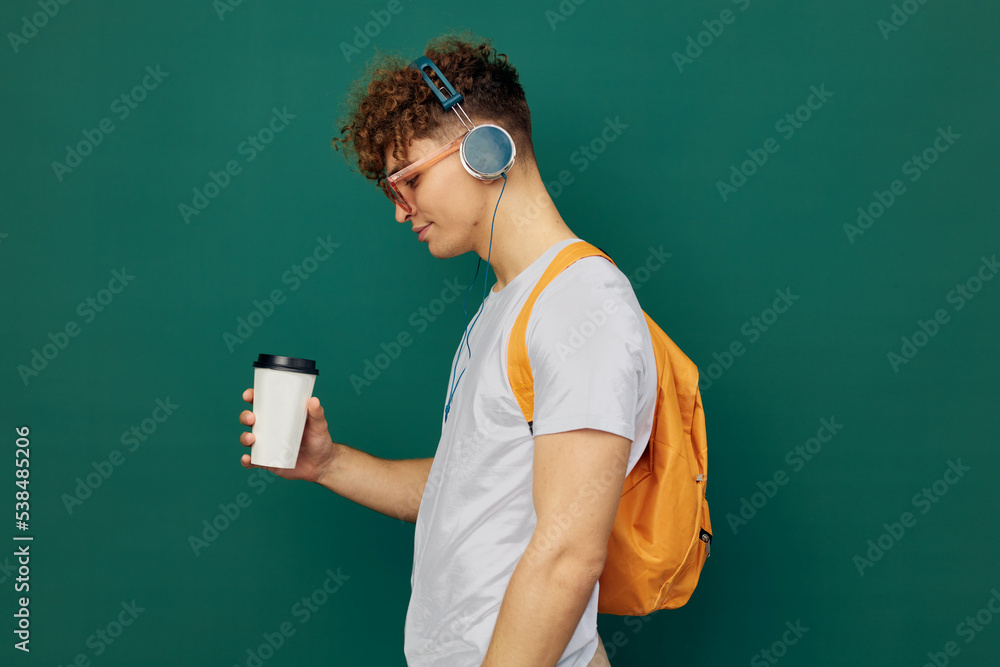 man stands on a green background listening to music with headphones with bright glasses and a backpack on his back holding a cardboard glass for hot drinks in his hand, standing sideways to the camera