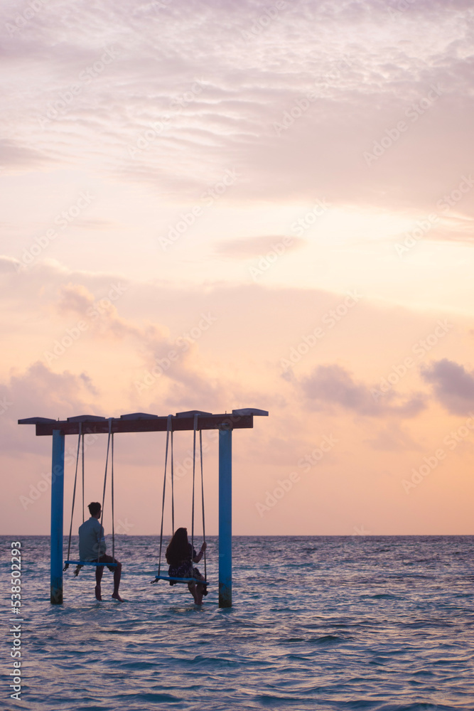 A picture of two couples sitting on a swing in the shallow sea of Maldives and looking at the beautiful pink sky during sunset