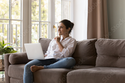 Pensive young Indian woman sit on couch with laptop, ponder over task studying on-line looks interested and focused, search solution, thinks internet purchase. Telework, modern tech usage for learning