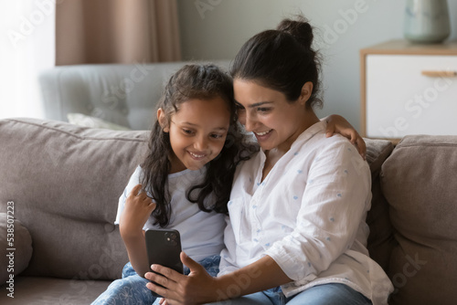 Attractive Indian woman and little cute daughter sit on sofa look at smartphone screen enjoy new mobile apps, calling through video call to family, spend time at home use cellphone. Modern tech, fun