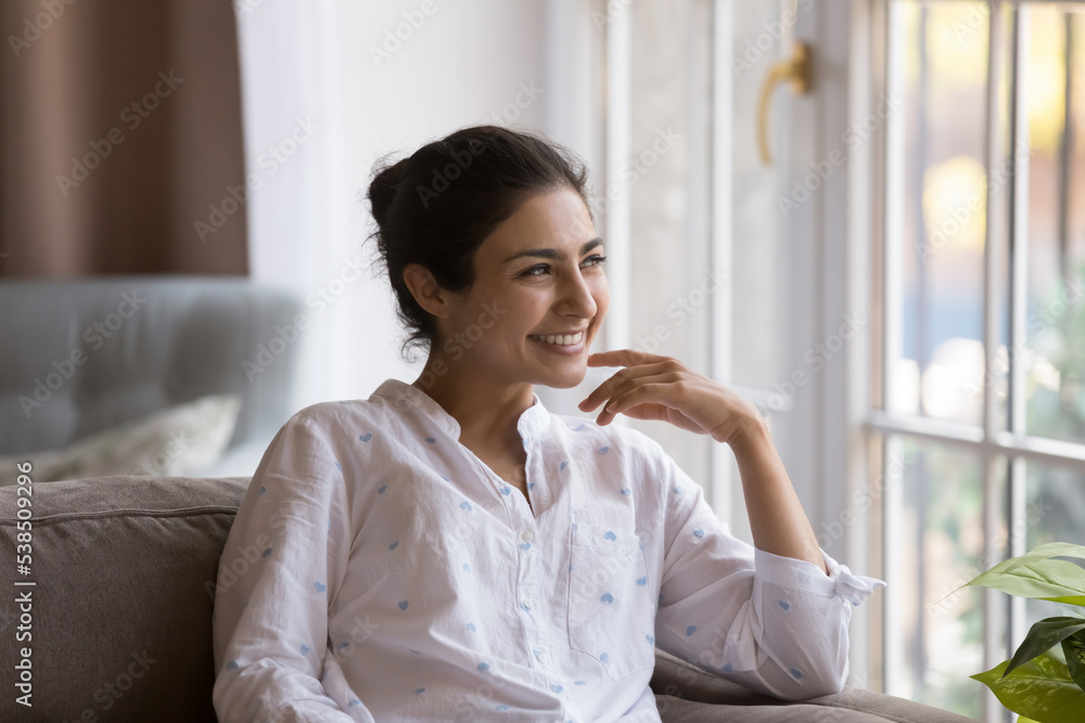 Young millennial dreamy Indian woman smile staring into distance while relaxing on cozy couch at modern home, enjoy daydreams and carefree weekend leisure feels happy, spend free time alone indoors Young millennial dreamy Indian woman smile staring into distance while relaxing on cozy couch at modern home, enjoy daydreams and carefree weekend leisure feels happy, spend free time alone indoors