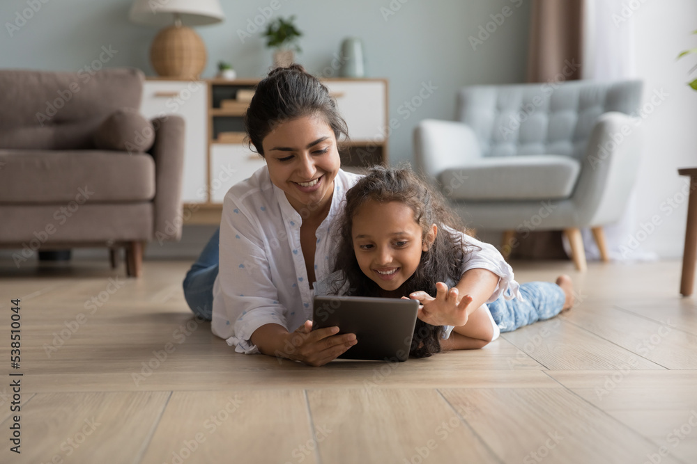 Educational application usage for preschooler children development, leisure on internet. Indian mother and little daughter lying on warm floor have fun use digital tablet, playing or buying on-line Educational application usage for preschooler children development, leisure on internet. Indian mother and little daughter lying on warm floor have fun use digital tablet, playing or buying on-line