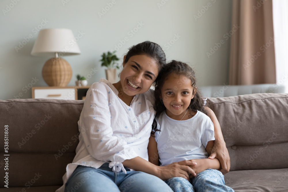 Portrait of happy Indian woman hugs little adorable daughter laughing seated together on sofa staring at camera enjoy pleasant communication through video conference app. Family bond, motherhood, fun Portrait of happy Indian woman hugs little adorable daughter laughing seated together on sofa staring at camera enjoy pleasant communication through video conference app. Family bond, motherhood, fun