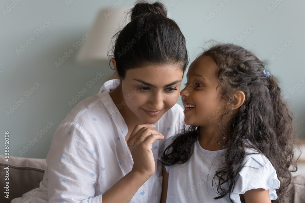 Close up of cute little Indian girl share secret on loving attentive mothers ear seated on sofa at home. Mom and daughter gossiping, have fun, enjoy communication and trustworthy relationships, trust