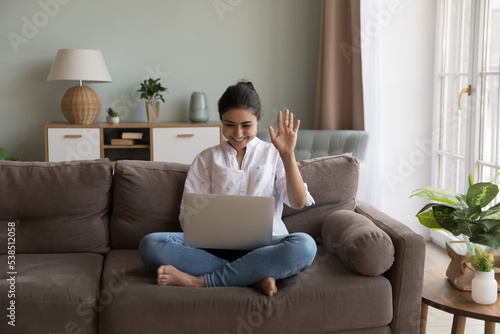 Happy Indian woman greeting family start video call seated on sofa with laptop, looks at device screen welcoming friend enjoy distancing communication using videoconference application and modern tech