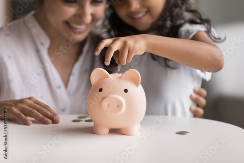 Smiling Indian mom and her little 6s cute daughter dropping coins into piggy bank. Loving parent teach child be thrifty, to save pocket money for future education or purchases, think about tomorrow