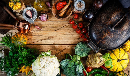 Autumn food background with organic farm vegetables: cauliflower, broccoli, root celery, pumpkin, herbs and spices on rustic wood kitchen table with cast iron pan, spice grinders, cutting board