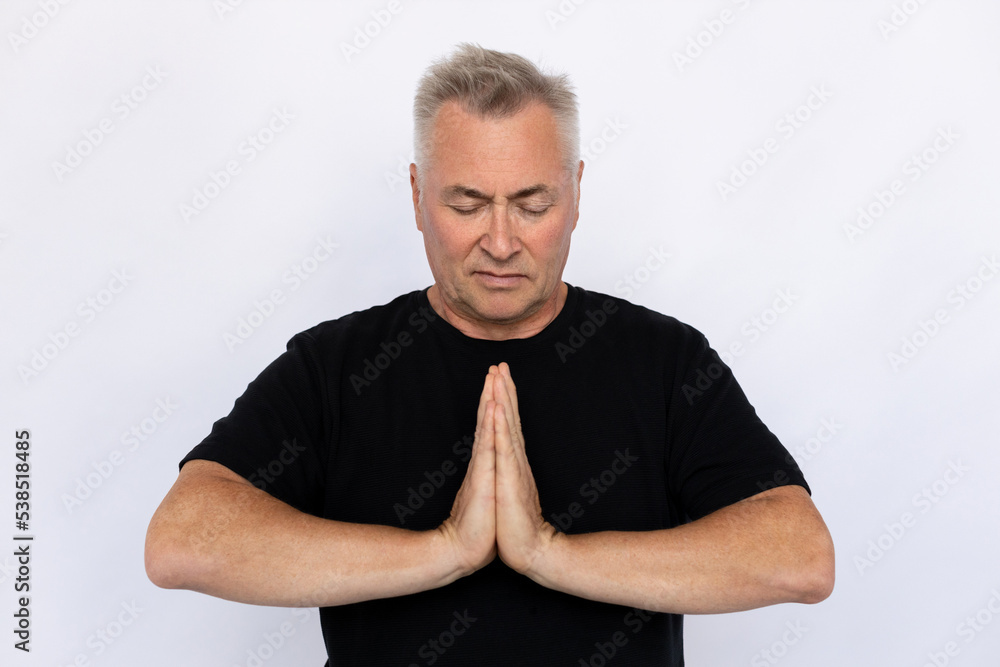 Senior man pressing hands together. Male model in black T-shirt clasping hands, standing with closed eyes, praying or meditating. Portrait, studio shot, meditation concept Senior man pressing hands together. Male model in black T-shirt clasping hands, standing with closed eyes, praying or meditating. Portrait, studio shot, meditation concept