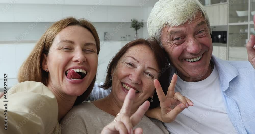 Close up 60s couple with adult daughter fooling in front of cellphone camera making funny faces showing V-sign have fun use modern tech. Blogging, multi-generational family leisure, video call concept Close up 60s couple with adult daughter fooling in front of cellphone camera making funny faces showing V-sign have fun use modern tech. Blogging, multi-generational family leisure, video call concept