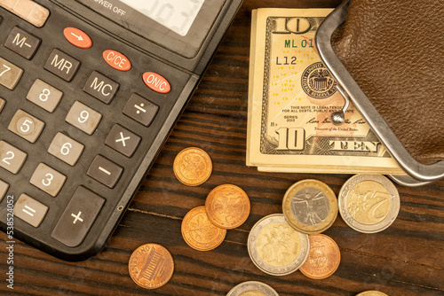 Dollar banknotes, euro coins, a vintage brown leather wallet and a calculator on a wooden surface.