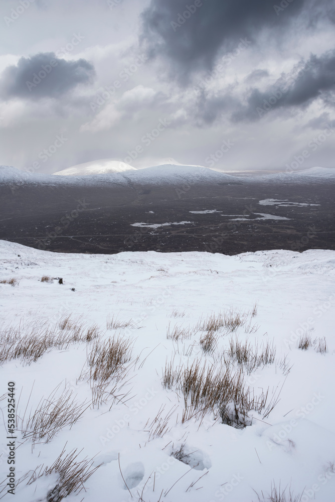 Epic Winter landscape image from mountain top in Scottish Highlands down towards Rannoch Moor during snow storm and spindrift off mountain top in high winds