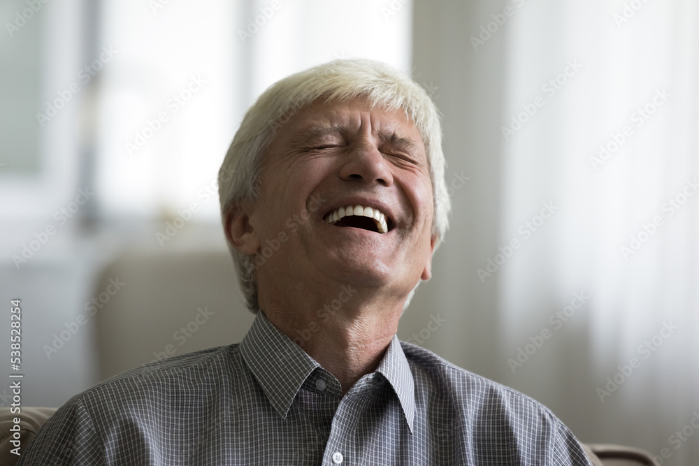 Close up shot, older man laughing at witty joke, happy senior giggling with eyes closed seated on sofa at home, spend weekend leisure watching amusing TV show, entertainment and fun, positive emotions Close up shot, older man laughing at witty joke, happy senior giggling with eyes closed seated on sofa at home, spend weekend leisure watching amusing TV show, entertainment and fun, positive emotions