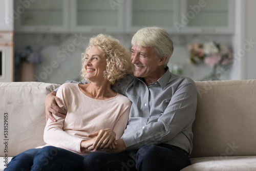 Elderly couple relaxing on sofa staring into distance. Serene attractive older wife, grey haired husband smile lookaside feeling carefree, enjoy calm retired life. Well-being family, happy marriage