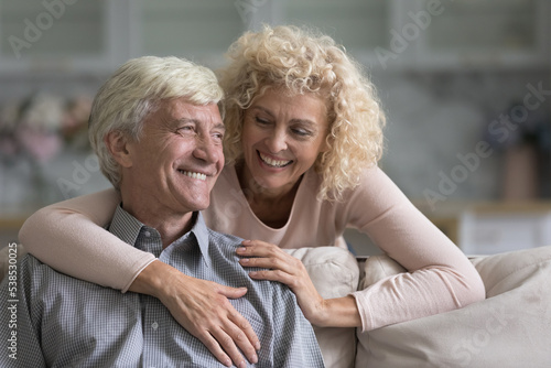 Happy well-being spouses, senior loving couple. Older smiling attractive wife hugging her beloved cheerful husband, spending time together at modern cozy home, enjoy leisure and harmonic relationship