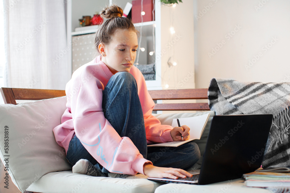 Millennial teen girl sit at couch in living room study on laptop making notes Millennial teen girl sit at couch in living room study on laptop making notes