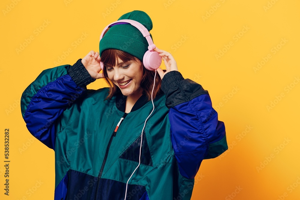 horizontal photo of a beautiful woman in a jacket and hat listening to music with pink headphones holding them with her hands horizontal photo of a beautiful woman in a jacket and hat listening to music with pink headphones holding them with her hands