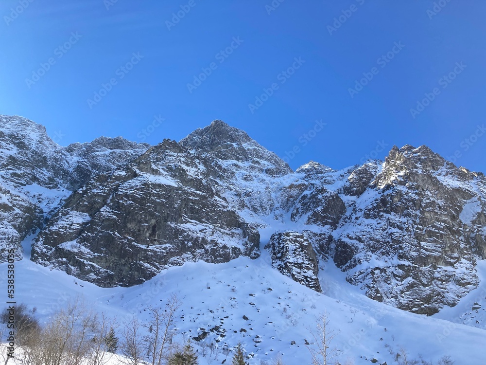 Winter mountains with snow in the winter rocky landscape. Winter nature landscape with mountains, trees, forest, plants and blue sky with clouds and sun. Hiking in the mountain with snowy path. Winter mountains with snow in the winter rocky landscape. Winter nature landscape with mountains, trees, forest, plants and blue sky with clouds and sun. Hiking in the mountain with snowy path.