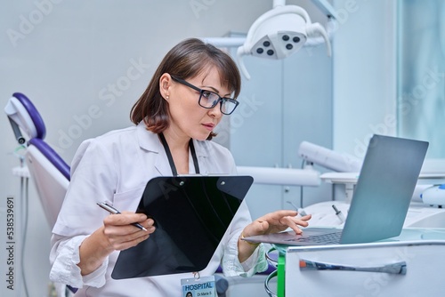 Doctor dentist working in office, using laptop, making notes on clipboard