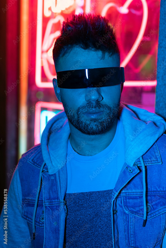 Hipster handsome man on the city streets being illuminated by neon signs. He is wearing leather biker jacket or asymmetric zip jacket with black cap, jeans and sunglasses Hipster handsome man on the city streets being illuminated by neon signs. He is wearing leather biker jacket or asymmetric zip jacket with black cap, jeans and sunglasses