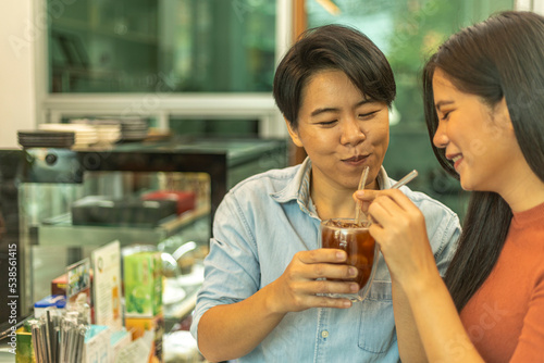 A young couple of lesbians holding a cup of ice coffee or tea together in a coffee shop. A lady is taking care her girlfriend with a cold drink. Two women share a cold coffee to each other.