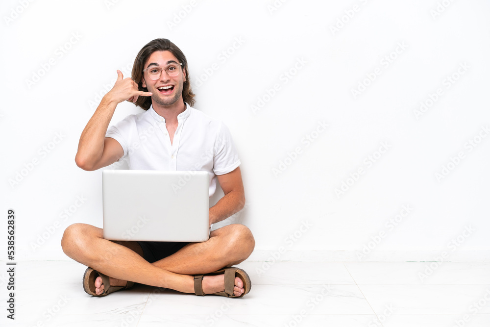 Young handsome man with a laptop sitting on the floor isolated on white background making phone gesture. Call me back sign Young handsome man with a laptop sitting on the floor isolated on white background making phone gesture. Call me back sign