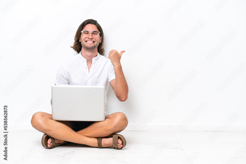 Young handsome man with a laptop sitting on the floor isolated on white background pointing to the side to present a product Young handsome man with a laptop sitting on the floor isolated on white background pointing to the side to present a product