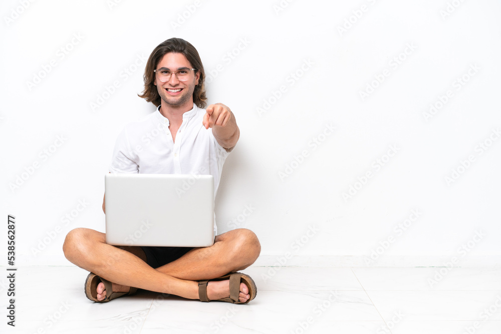 Young handsome man with a laptop sitting on the floor isolated on white background points finger at you with a confident expression Young handsome man with a laptop sitting on the floor isolated on white background points finger at you with a confident expression