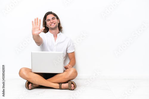 Young handsome man with a laptop sitting on the floor isolated on white background counting five with fingers