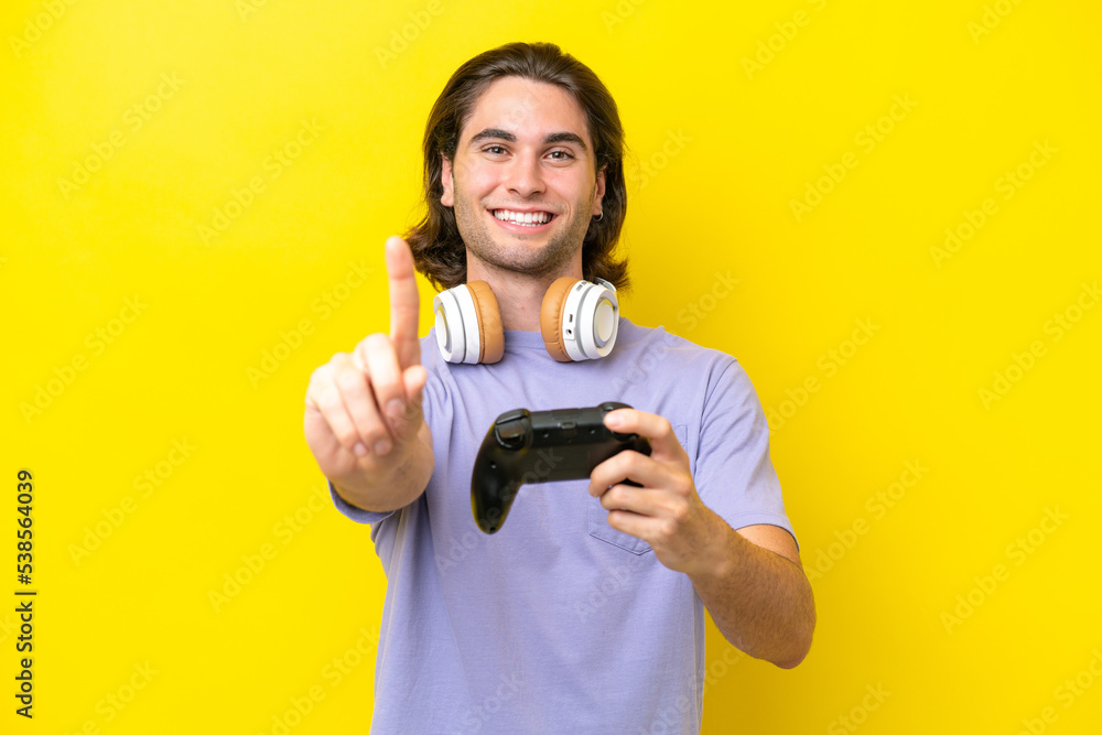 Young handsome caucasian man playing with a video game controller over isolated on yellow background showing and lifting a finger Young handsome caucasian man playing with a video game controller over isolated on yellow background showing and lifting a finger