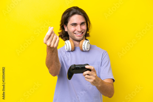 Young handsome caucasian man playing with a video game controller over isolated on yellow background making money gesture
