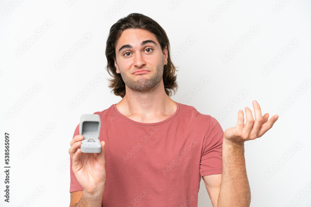Young handsome man holding a engagement ring isolated on white background making doubts gesture while lifting the shoulders Young handsome man holding a engagement ring isolated on white background making doubts gesture while lifting the shoulders