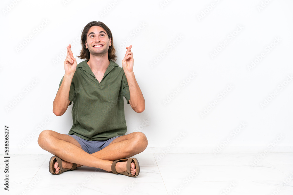 Young handsome man sitting on the floor isolated on white background with fingers crossing Young handsome man sitting on the floor isolated on white background with fingers crossing