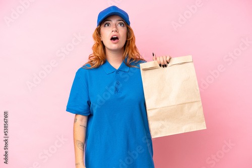 Young caucasian woman taking a bag of takeaway food isolated on pink background looking up and with surprised expression