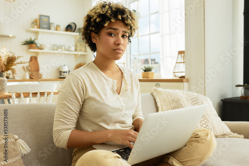 Upset tired exhausted african america young female translator working at home as freelancer sitting on couch with laptop on knees looking at camera with sad pitiful facial expression